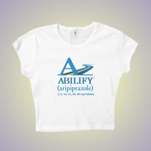 May include: White crop top featuring the word "ABILIFY" in blue, with a stylized road design. Below, the text "(aripiprazole)" and tablet dosages are printed in blue. The shirt is a casual style.