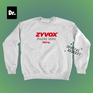 Puede incluir: Una sudadera gris claro con el texto rojo "ZYVOX (linezolid) comprimidos 600 mg". Las palabras "SPECIAL REQUEST" están impresas en negro en la manga derecha. Un cuadrado negro con "Dr." en blanco está en la esquina superior izquierda.