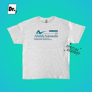 May include: A light gray heathered t-shirt featuring a blue and black graphic print on the chest. The text reads Abilify Asimtufii (aripiprazole) extended release suspension for injection, Every Two Months, 960mg. A Dr. logo and Special Request text appear.