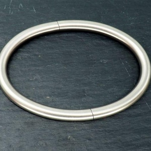 May include: A silver-colored, circular piece of jewelry, possibly a necklace or bracelet, with a seamless design. The metal appears to be polished, reflecting light. The jewelry is set against a dark, textured background.