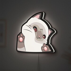 May include: A wall decoration in the shape of a cartoon cat. The cat is white with grey details, pink ears and paw pads, and brown eyes. It has a black outline and is mounted on a dark grey wall.