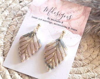 May include: A pair of leaf-shaped earrings with gold-colored accents. The earrings have a pink, gray, and white marbled design. The earrings are displayed on a white card with the text "Mdesigns" and "Laser cut earrings, accessories & more".