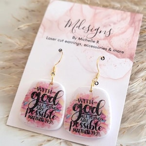 May include: A pair of laser-cut earrings with gold-colored hooks. Each earring features a white background with a watercolor design in pink, yellow, and blue. The text "With God all things are possible Matthew 19:26" is printed in black.