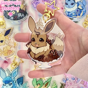 Puede incluir: Un llavero acrílico transparente con un personaje de dibujos animados marrón con orejas grandes, una cola esponjosa y un pecho color crema. El personaje está en un cuenco de helado con salsa de chocolate y crema batida. El llavero tiene un cierre dorado en forma de estrella.