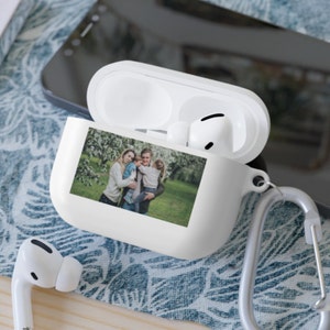 May include: White Airpod case with a photo of a family of three in a park. The photo shows a woman, a man, and a young child.