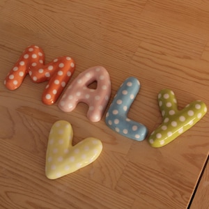May include: A set of five ceramic letters spelling the word MALY, with an additional heart-shaped piece. Each piece features a glossy finish in various pastel colors including orange, pink, blue, green, and yellow, all decorated with white polka dots.