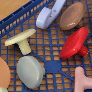 Può includere: Una collezione di pomelli e ganci in ceramica in vari colori, tra cui rosso, giallo, azzurro, pesca, grigio, marrone e verde. Gli oggetti hanno superfici lisce e arrotondate e sono disposti su un vassoio di plastica blu a griglia.