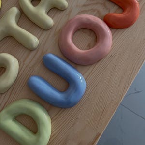 Può includere: Una collezione di lettere e forme in ceramica tridimensionali, arrotondate e lucide, in colori pastello tra cui giallo pallido, rosa tenue, azzurro e arancione, disposte su una superficie in legno chiaro con venature visibili.
