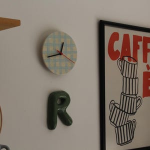Puede incluir: Un reloj de pared redondo con patrón de cuadros azul claro y blanco, manecillas negras y segundero rojo. Debajo hay una letra R de cerámica verde oscuro. A la derecha, un póster enmarcado con el texto rojo CAFFÈ y tazas a rayas.