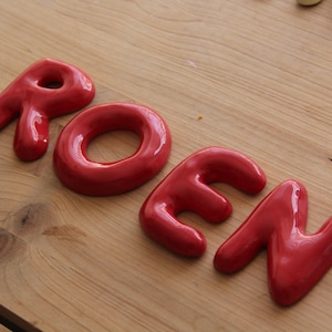 Könnte beinhalten: Vier einzelne glänzende rote Keramikbuchstaben, die das Wort ROEN bilden. Jeder Buchstabe hat ein glattes, abgerundetes und aufgeblähtes Aussehen mit einer reflektierenden Oberfläche. Die Buchstaben sind in einer Reihe auf hellem Holz angeordnet.