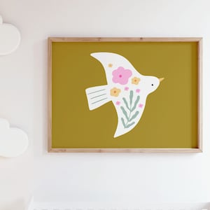 Op de afbeelding: Een witte vogel met roze en gele bloemen vliegt tegen een mosterdgele achtergrond. De vogel zit in een houten lijst en hangt aan een witte muur.