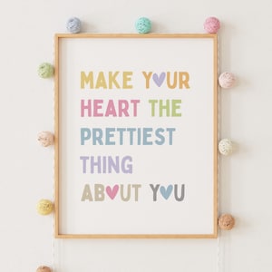 Könnte beinhalten: Ein gerahmter Druck mit dem Text "Make your heart the prettiest thing about you" in Regenbogenfarben mit Herzen. Der Druck ist von einer Schnur mit bunten Wollkugeln umgeben.