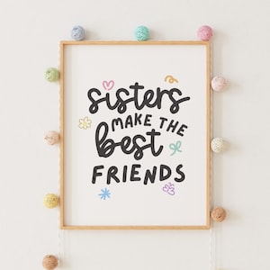 Może przedstawiać: Wydruk w ramie z tekstem "Sisters make the best friends" w czarnym, ozdobnym piśmie. Wydruk jest otoczony jasną drewnianą ramą i ozdobiony kolorowymi kulkami z włóczki.