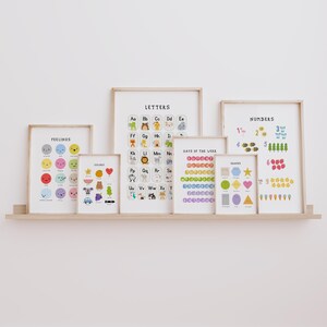 May include: A collection of framed educational posters displayed on a wooden shelf. The posters feature colorful illustrations of letters, numbers, shapes, colors, days of the week, and feelings. The frames are a light wood color.