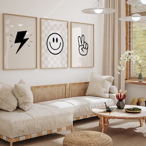 Könnte beinhalten: Ein weißes Sofa mit drei Kissen und einem Holzrahmen steht in einem Wohnzimmer mit einem Couchtisch und einem geflochtenen Hocker. Drei gerahmte Drucke mit Schwarzweißillustrationen eines Blitzes, eines Smiley-Gesichts und eines Peace-Zeichens hängen an der Wand.