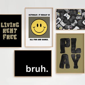 May include: Five framed art prints with various designs. One black print reads "LIVING RENT FREE" in tan letters. Another features a yellow smiley face with the text "ALL FUN AND GAMES." A third print displays a pattern of video game controllers.