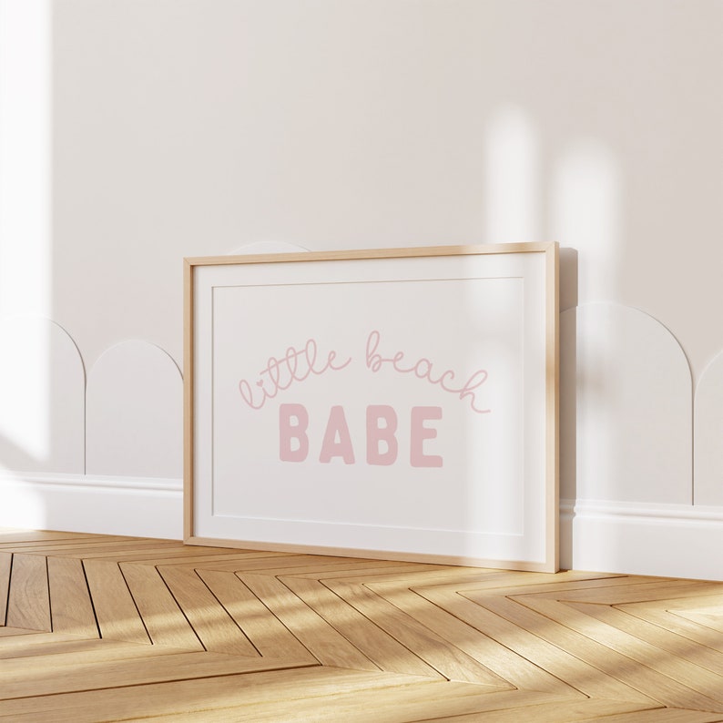 May include: A framed print with a white background and a pink inscription that reads "little beach BABE".