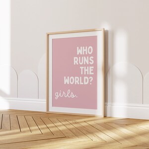 May include: A framed pink poster with the text "Who runs the world? girls." in white lettering.