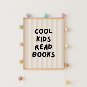 May include: A framed print with a white and beige striped background. The text "COOL KIDS READ BOOKS" is written in black on the print. The print is surrounded by a string of colorful pom-poms.