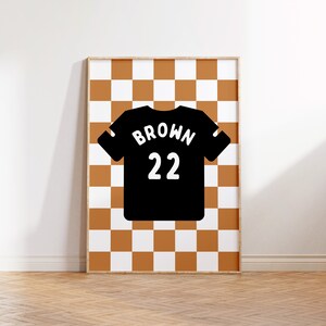 Op de afbeelding: Een ingelijst print met een zwart-wit geruit patroon. Een zwart shirt met het woord "BROWN" en het nummer "22" in wit is gecentreerd op het geruite patroon.