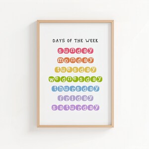 May include: A framed print with the text "DAYS OF THE WEEK" in black. The days of the week are listed in a rainbow of colors: red, orange, yellow, green, blue, purple. The frame is a light brown color.