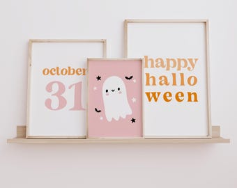 May include: Three framed Halloween-themed prints on a wooden shelf. The prints feature the words "october 31" in orange and pink, a cute ghost illustration on a pink background, and the words "happy halloween" in orange. The frames are light-colored.