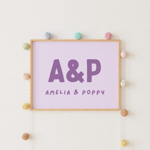 Puede incluir: Una impresión enmarcada con un fondo morado claro y el texto "A&P Amelia & Poppy" en morado oscuro. El marco es de madera y tiene un color marrón claro. La impresión está rodeada de una cuerda de pompones coloridos.