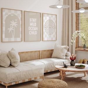 Puede incluir: Un sofá beige claro con dos almohadas y un marco de madera. El sofá está en una sala de estar con una mesa de café, un pequeño taburete y tres impresiones enmarcadas en la pared. Las impresiones muestran una palmera, el texto "Stay Wild Ocean Child" y un sol poniéndose sobre el océano.