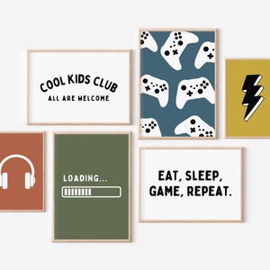 May include: A collection of framed wall art with a gaming theme. The art includes a white background with the text "COOL KIDS CLUB ALL ARE WELCOME", a blue background with white game controllers, and a yellow background with a black lightning bolt.