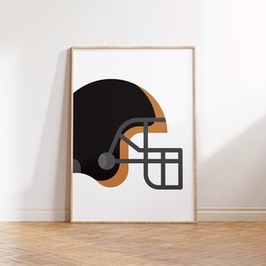 Op de afbeelding: Een zwarte en bruine voetbalhelm met een grijs gezichtsmasker. De helm staat op een witte achtergrond.