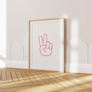 Op de afbeelding: Een ingelijst print met een roze vredesteken op een witte achtergrond. De lijst is van licht hout en de print hangt aan een witte muur.