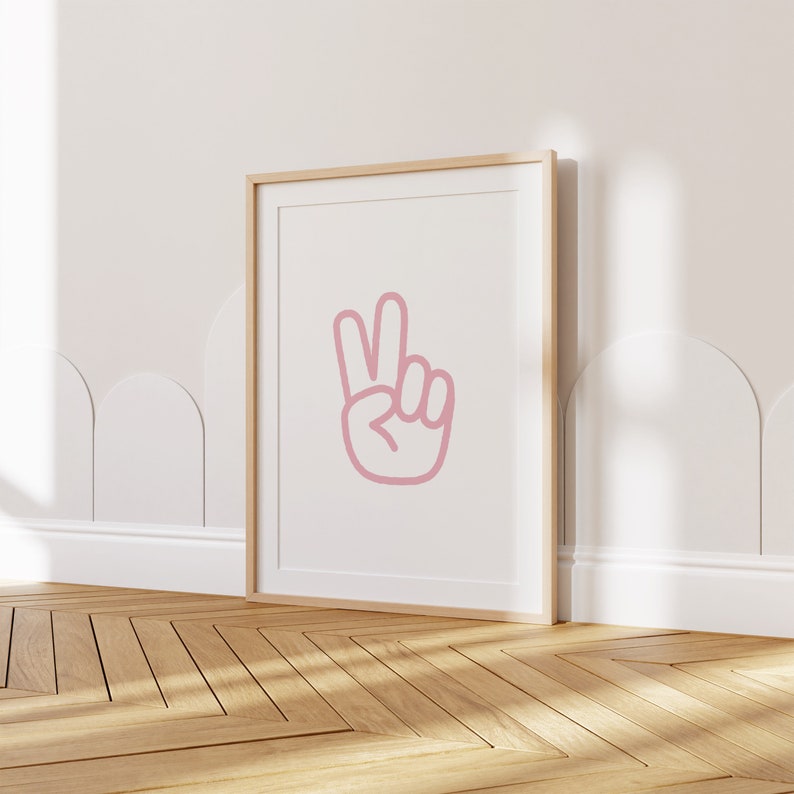Op de afbeelding: Een ingelijst print met een roze vredesteken op een witte achtergrond. De lijst is van licht hout en de print hangt aan een witte muur.