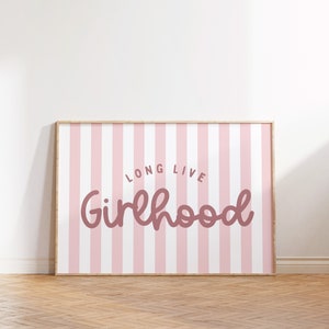 Puede incluir: Un estampado enmarcado con un fondo de rayas rosas y blancas. Las palabras "LONG LIVE Girlhood" están escritas en una fuente cursiva en un color rosa más oscuro. El marco es de color madera clara. El estampado se muestra sobre un suelo de madera.