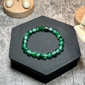 May include: A green gemstone bracelet with a silver magnetic clasp. The bracelet is made of round beads and is displayed on a black hexagonal stand.