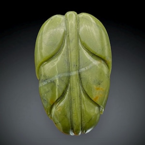 Puede incluir: Un colgante de hoja de piedra verde tallada. El colgante tiene un diseño detallado con vetas y un acabado pulido. La piedra tiene variaciones de color, con vetas blancas y amarillas. La forma general es simétrica y alargada.