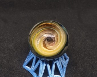 May include: A glass marble with a swirling pattern of black, white, and gold. The marble is sitting on a blue plastic stand.