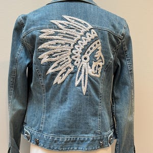 May include: Pale blue denim jacket featuring a white embroidered Native American headdress on the back. The jacket has a classic collar, button fastenings, and silver-tone rivets. A stylish and unique outerwear piece.