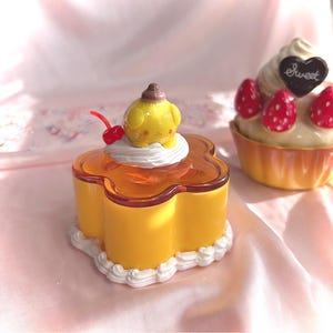 May include: A yellow flower-shaped container with a translucent orange lid, topped with a small yellow character figure, white whipped cream, and a red cherry. Beside it sits a cupcake-shaped container with cream, red strawberries, and a heart.