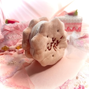 May include: A plush, light brown cookie-shaped coin purse with the embroidered words "Eat me". The purse has a white zipper and is surrounded by other pink, decorative items, including a small cake and a pink bow.