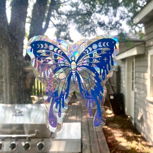 May include: A holographic sticker of a blue butterfly with a moon and star design. The butterfly has dripping paint accents and iridescent wings.