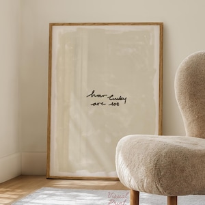 May include: A framed print with the text "how lucky are we" in black script. The print is displayed in a light-colored room with a beige, upholstered chair and a light gray rug. A stack of books and a small bowl are on the floor.