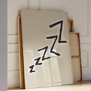 May include: A framed artwork with a beige background and a gold frame leans against a wall. The artwork features the word "zzz" in black, stylized lettering. The image is set in a room with a wooden floor and white walls.