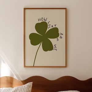 May include: A framed print with a green four-leaf clover and the text "HOW LUCKY ARE WE" in black. The artwork is set against a cream background and framed in a wooden frame. The print is displayed above a bed with striped bedding.