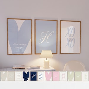 May include: Three framed art prints with wooden frames. The prints feature a blue heart, the letter "H", and "XO XO" in a light blue colour. The text "How lucky are we" is also visible. A lamp and books are on a white table below the prints.