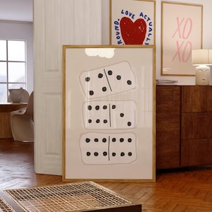 Op de afbeelding: Een ingelijste print met drie dominostenen met zwarte stippen op een beige achtergrond. Twee andere ingelijste prints zijn zichtbaar, een met een rood hart en de woorden "LOVE ACTUALLY ROUND" en een andere met "XO XO" in roze.