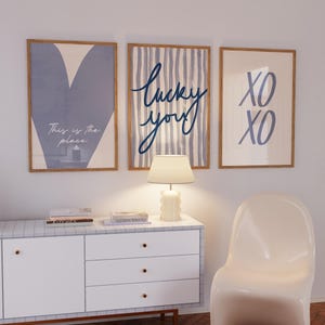 Pode incluir: Três impressões emolduradas com designs minimalistas. As impressões apresentam um coração azul, a frase "lucky you" em escrita e "XO XO" em azul. As molduras são de cor castanho claro, e as impressões são exibidas acima de um armário branco.