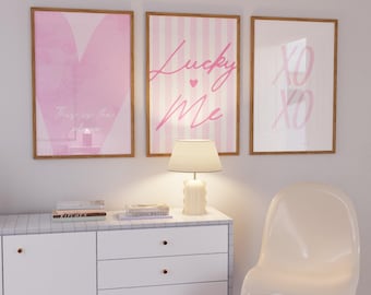 Könnte beinhalten: Drei gerahmte Kunstdrucke mit rosa Designs. Der linke Druck zeigt ein rosa Herz mit dem Text "This is the place". Der mittlere Druck hat rosa Streifen und die Worte "Lucky Me". Der rechte Druck zeigt "XO XO". Die Rahmen sind hellbraun.