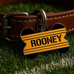May include: A dog tag in the shape of a bone, with the name "ROONEY" in bold yellow letters on a black and yellow striped background. The tag is attached to a brown leather dog collar.