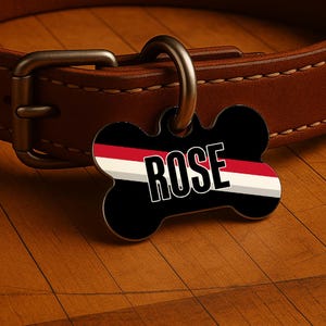 May include: A black, bone-shaped pet tag with the name "ROSE" in white, outlined letters. The tag features a red and white stripe design and is attached to a brown leather dog collar with a metal clasp.