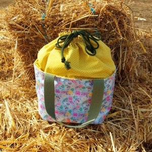 May include: A yellow and white drawstring bag with green straps and a floral print. The bag is sitting on a pile of hay.